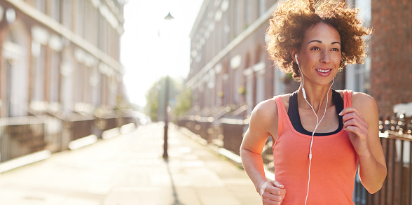 a young woman running in a street on her morning or evening run . She is backlit by a rising low sun , and is pictured staring ahead with determination , before heading off on her morning jog.She is wearing a vest top , trainers and leggings , and earphones. The sun is rising at dawn behind her or setting at sunset . Lens flare streaks across the frame .