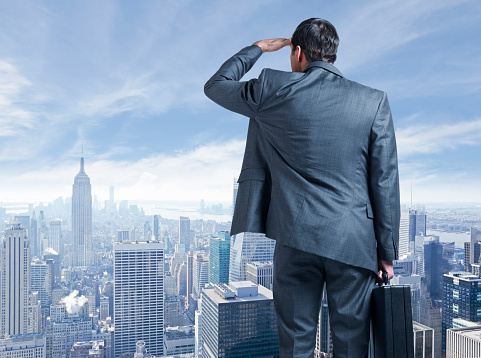 A businessman carries his briefcase and places his other hand over his brow as he looks out towards the New York City skyline.