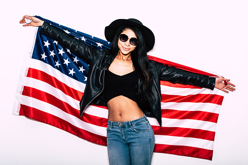 Beautiful young mixed race woman carrying American flag and smiling while standing against white background