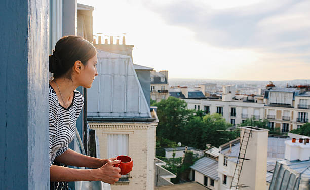 Young woman enjoying the view from a Parisian apartment Vintage toned image of a young woman relaxing, drinking coffee, on the small balcony - window of her beautiful apartment on Montmartre, Paris. Taken in the magic hour just as the sun sets down over Parisian city scape in the background and the sunrays paint her hair bright. ile de france stock pictures, royalty-free photos & images