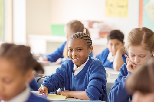 A female Elementary student of African decent,  sits at her desk in class as she works on a writing assignment.  She is dressed casually and is looking up from her books to smile.   Her peers can be seen seated around her in the background at other desks, working away independently as well.