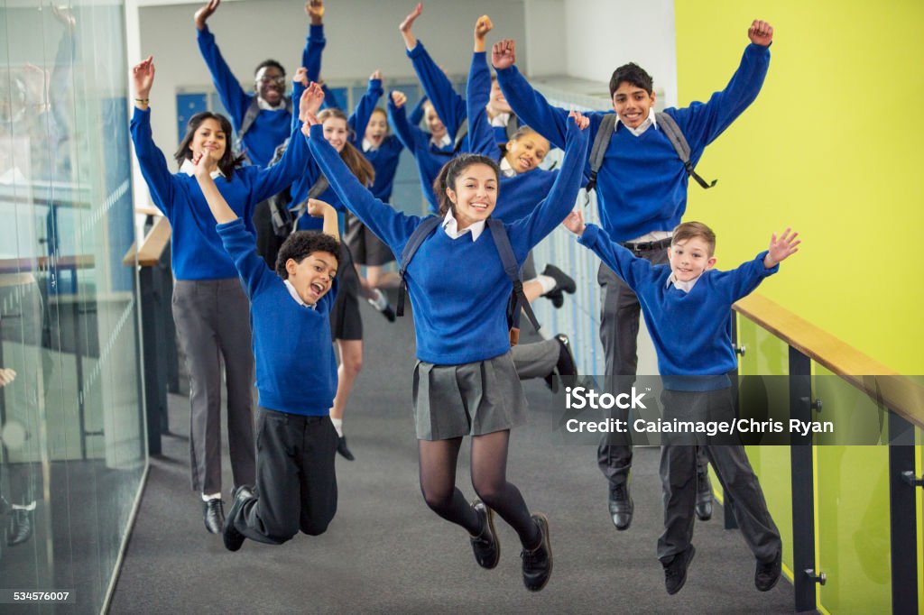 Enthusiastic high school students wearing school uniforms smiling and jumping in school corridor  School Building Stock Photo