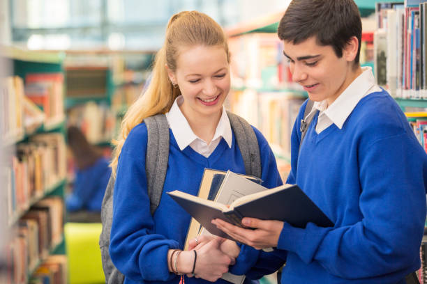 istock Two smiling high school students wearing school uniforms looking at book in library 534576133