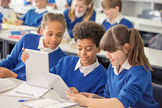 grundschulkinder in blauen schuluniformen mit digitalen tablets am schreibtisch im klassenzimmer - schulkind stock-fotos und bilder