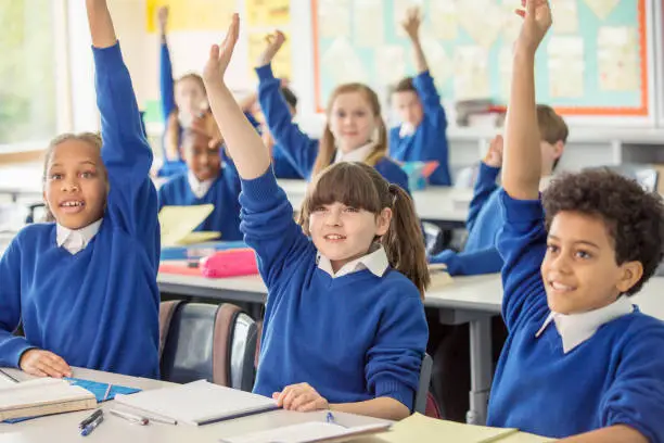 Photo of Elementary school children wearing blue school uniforms raising hands in classroom