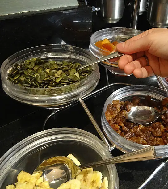 Photo showing a series of glass dishes / bowls filled with a selection of dried fruit, nuts and seeds, including pumpkin seeds, sultanas, dried banana slices and dried apricots.  The seeds and dried fruit are part of a hotel breakfast buffet, where guests help themselves and sprinkle the seeds / nuts on top of their breakfast cereal, muesli, yogurt / yoghurt or hot porridge.  A hand is pictured with a teaspoon, scooping up a spoonful of pumpkin seeds.