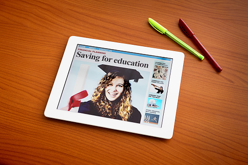 The screen of a digital tablet shows a newspaper front page with a pretty, smiling graduate and the headline: Saving ror education. Copy space on the wooden desk background.