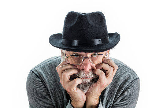 A curious senior adult man wearing a black fedora hat is looking up over his glasses with his chin on his hands while leaning forward toward the camera.