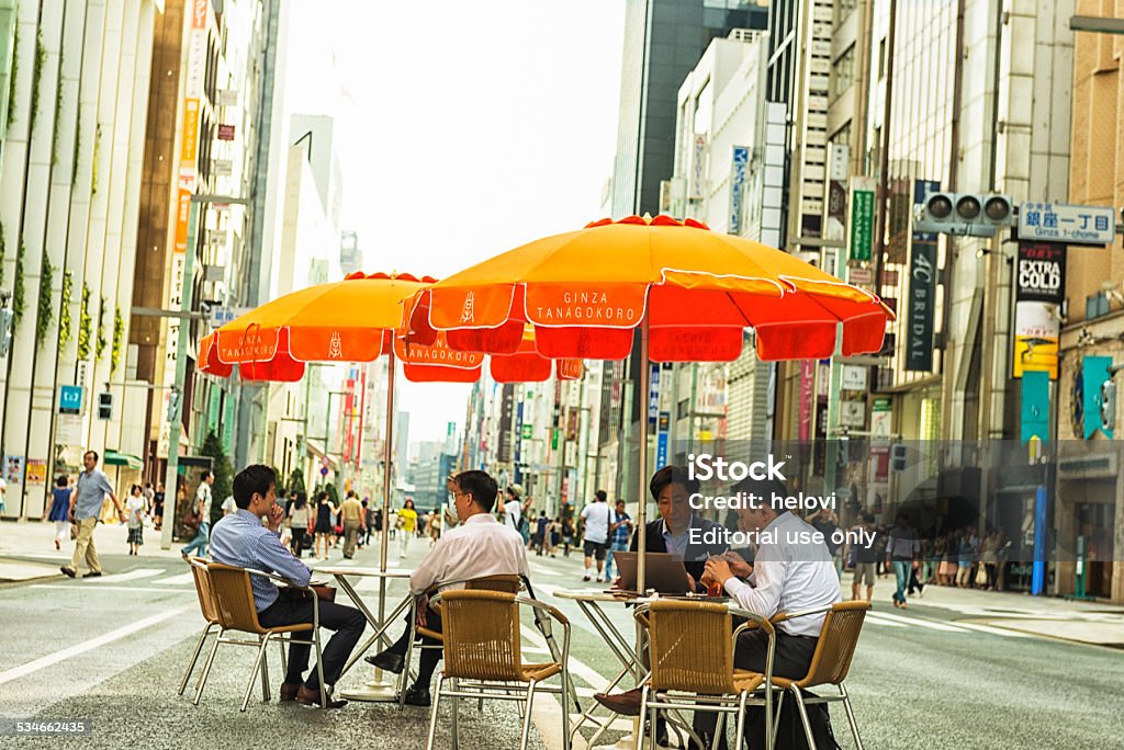 Ginza Shopping District Tokyo, Japan - August 17, 2014: Ginza Shopping District.  Each sunday the main street in Ginza is closed for trafic, than people sit at the tables under parasol in the middle of the street, meeting friends or just resting after their shopping. Here some bussinesman sit at the table, looking at their laptop, all around the street there are famous shops.  Ginza Stock Photo