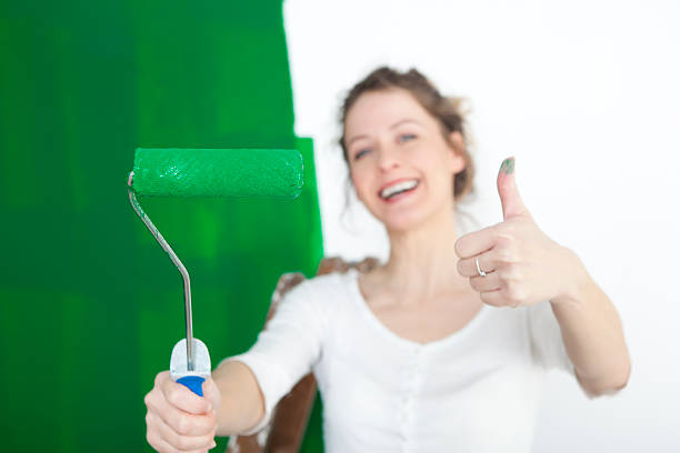 a woman painting a wall green and holds the paint roller to the camera