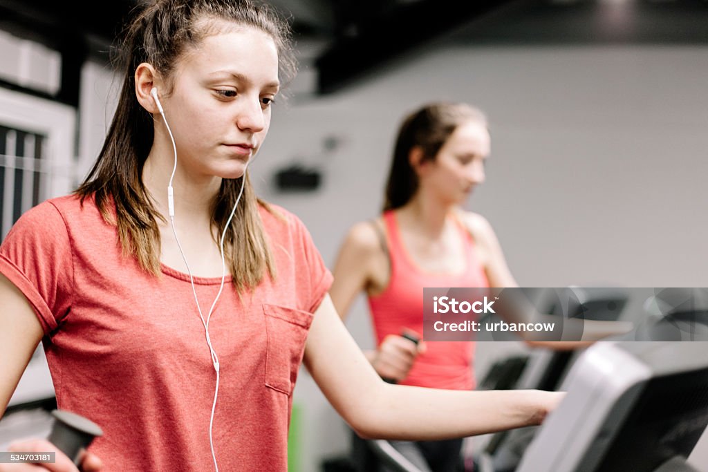 Young women using elliptical trainers, wearing earphones Two young women using cross training machines, wearing earphones and listening to music 2015 Stock Photo