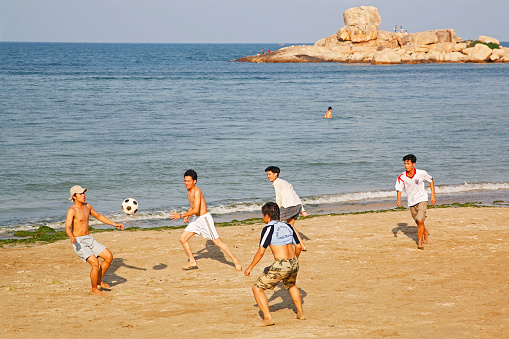 Nha Trang, Vietnam - March 27, 2006: A group of young men work up quite a sweat playing a friendly game of football (soccer) late one afternoon on a scenic beach in Nha Trang.  In the background people clamber over some of the spectacular rocks in Nha Trang Harbour.  Like many Asian countries, the Vietnamese love their round-ball football.  