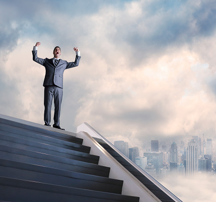 A businessman with his arms raised in victory, stands at the top of a stairway. A big city skyline in the background is partially obscured by fog and clouds. The camera is positioned below the stairway and looks up towards the businessman.  A large are of negative space exists in the clouds allowing for copy.