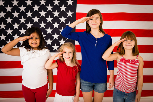 Color photo of four happy, patriotic, young girls saluting to honor America and our troops in front of a large American flag. Differing ethnicity portraying unity, friendship, acceptance and peace.