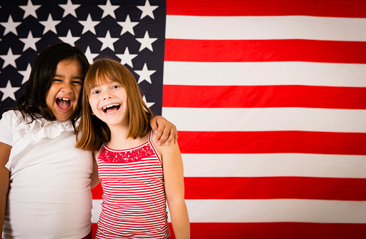 Color photo of two happy, patriotic, young girls laughing together with arms around each other in front of a large American flag. Differing ethnicity portraying unity, friendship, acceptance and peace.