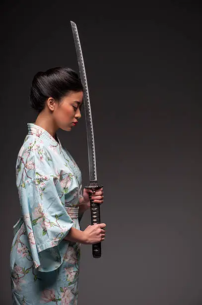 Total concentration. Side view portrait of young beautiful Japanese woman in kimono holding katana sword by her head while standing against grey background