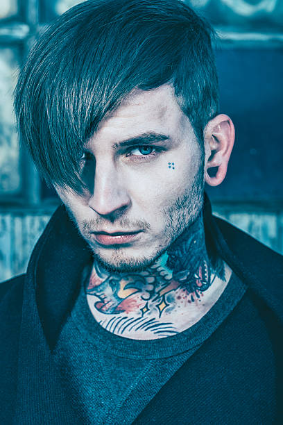 Headshot of a man in his 20s. He has funky hair and looks at the camera. Some of the hair covers on of his eyes. He has tattoos on both neck and face.