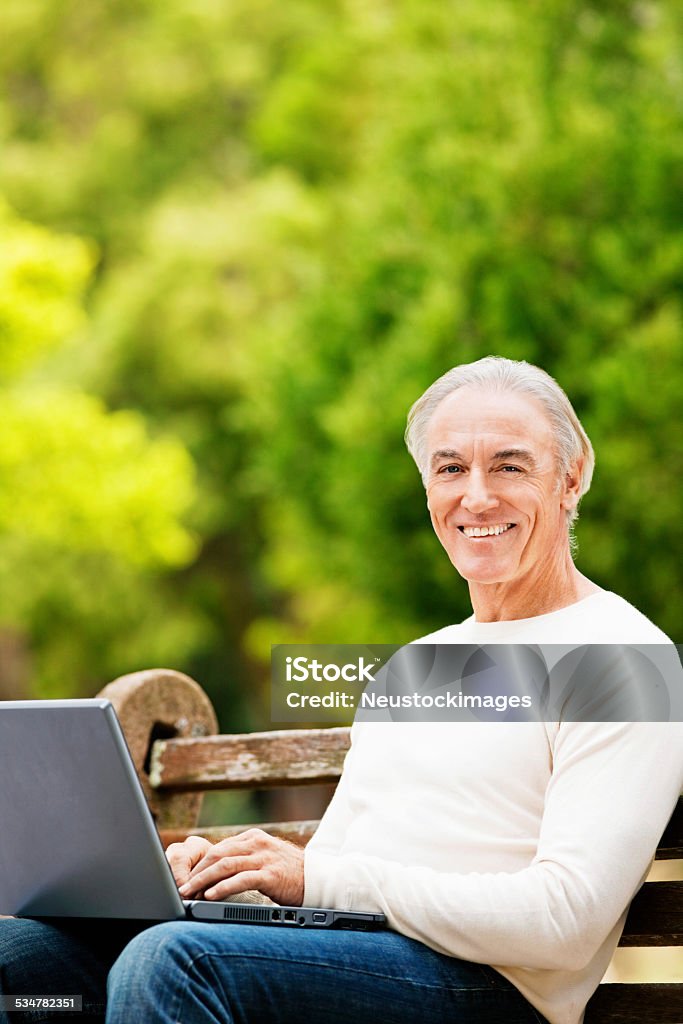 Senior Man Sitting on Park Bench With Laptop A senior man is smiling and sitting on a park bench with a laptop. Vertical shot. 2015 Stock Photo