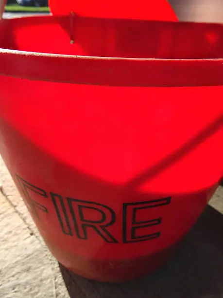 Photo showing a red plastic fire bucket filled with sand.  This bucket is located at a fuel filling station, so that any drivers who spill petrol / gas or diesel whilst filling their cars can soak up the spillage with the sand - preventing a potential fire hazard.  The bucket is bright red in colour, as this is the universal colour of fire-fighting equipment and fire extinguishers - and its red colour also makes it stand out on the filling station forecourt.