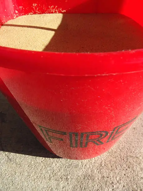 Photo showing a red plastic fire bucket filled with sand.  This bucket is located at a fuel filling station, so that any drivers who spill petrol / gas or diesel whilst filling their cars can soak up the spillage with the sand - preventing a potential fire hazard.  The bucket is bright red in colour, as this is the universal colour of fire-fighting equipment and fire extinguishers - and its red colour also makes it stand out on the filling station forecourt.