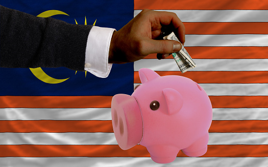 Man putting dollar into piggy rich bank national flag of malaysia in foreign currency because of inflation