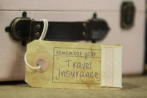 Paper tag with the handwritten words 'remember your travel insurance' on vintage suitcase.