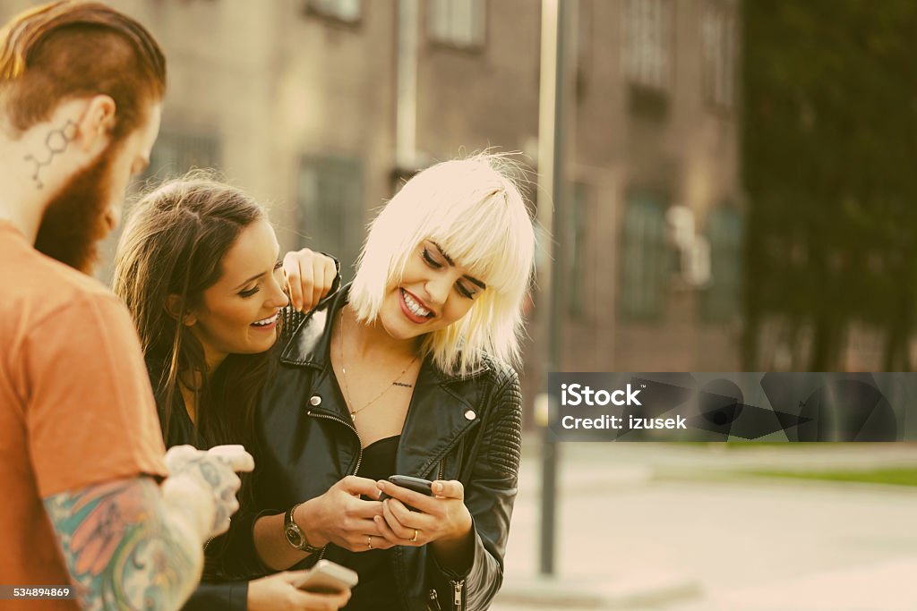 Two young woman using smart phones outdoor Outdoor portrait of two happy friends holding smart phones in hands. Bearded man with tatoo standing in the foreground. Blond Hair Stock Photo