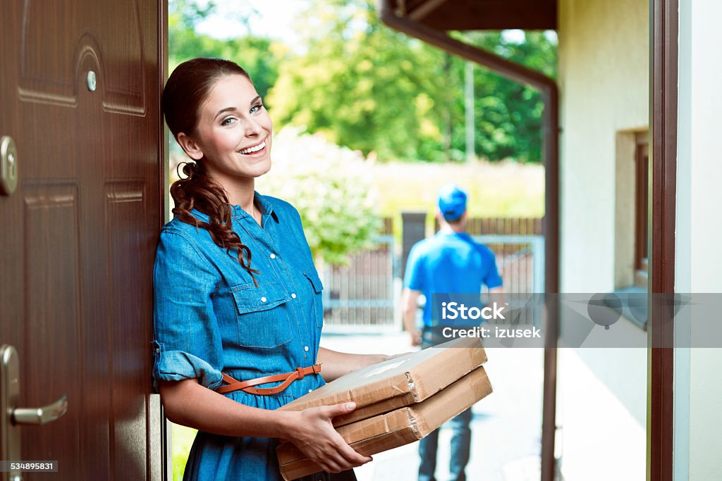 Young woman holding packages Young woman standing at the entrance door and holding parcels in hands, smiling at camera. Delivery man in the background. 2015 Stock Photo