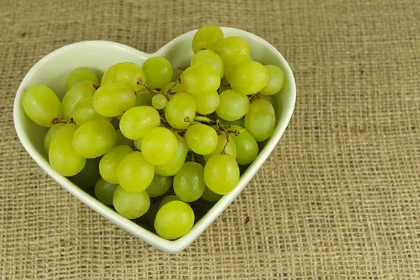 Green grapes in a white heart shaped bowl on a rustic hemp background.