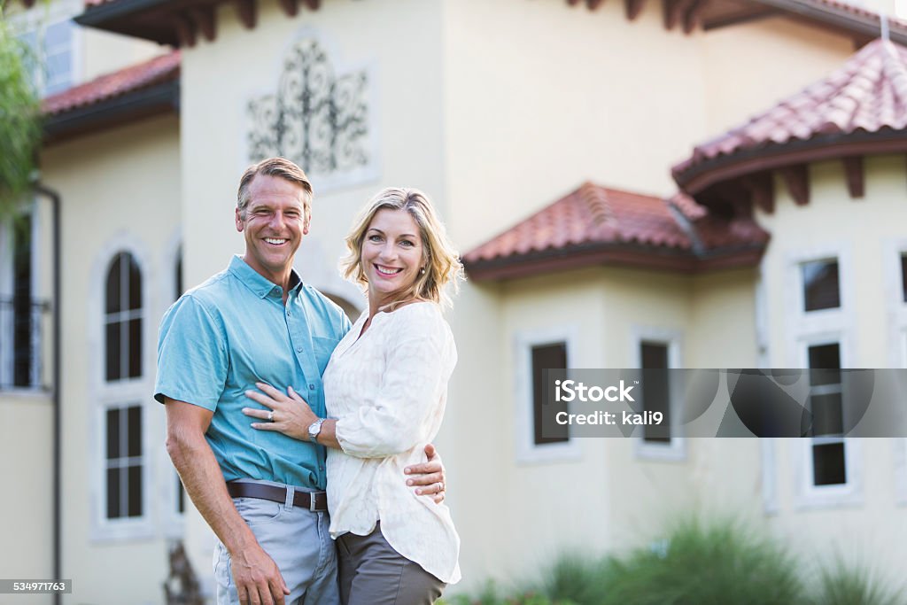 Mature couple standing in front of their mansion Affluent mature couple standing in front of their dream home, a huge mansion.  They are standing with arms around each other, smiling at the camera. Couple - Relationship Stock Photo