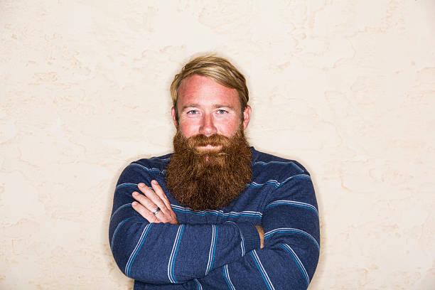 Handsome man with blond hair and a long red beard, standing with arms folded.