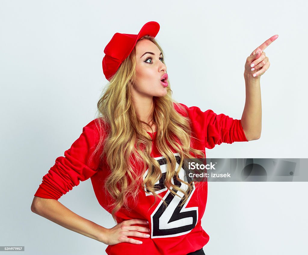 Surprised young woman pointing at copy space Side view of excited young woman wearing red sport blouse and cap pointing at copy space. Studio shot, white background. 16-17 Years Stock Photo