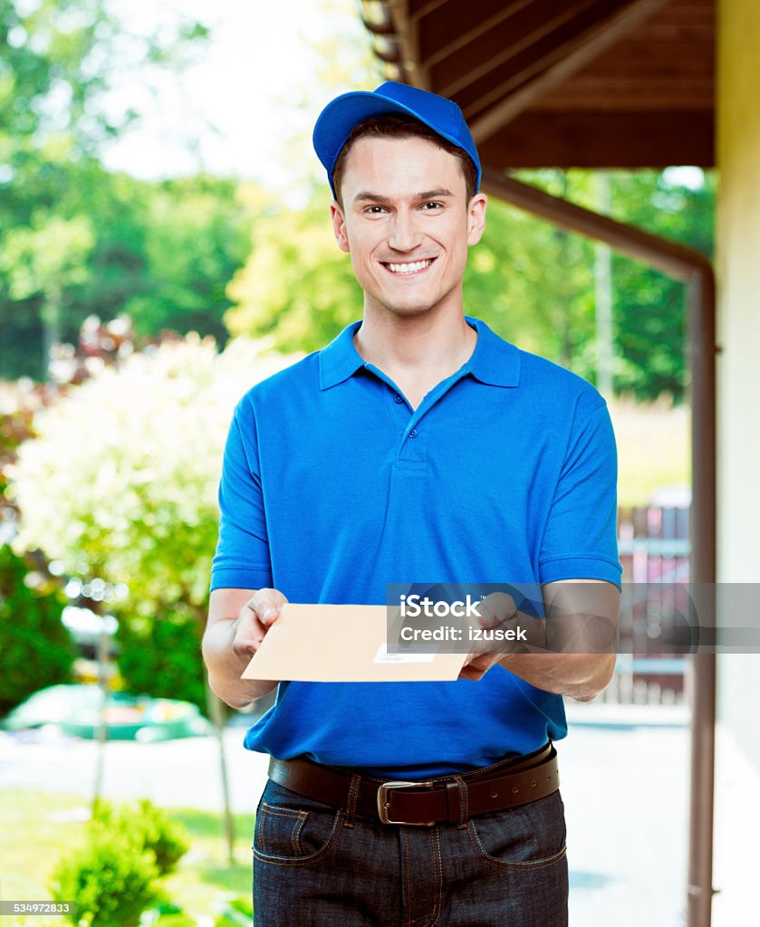 Postman delivering a mail Portrait of young postman standing outdoor, holding an envelope in hands and smiling at camera. Delivering Stock Photo