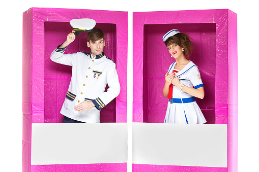Young beautiful boy and girl looking like dolls dressed in marine style costumes standing in big pink boxes