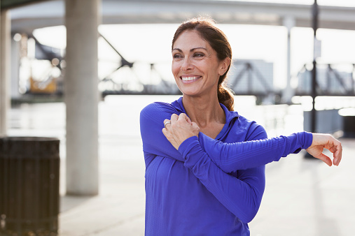 Mature woman taking a break from running to stretch her arms, looking at the view on the waterfront.