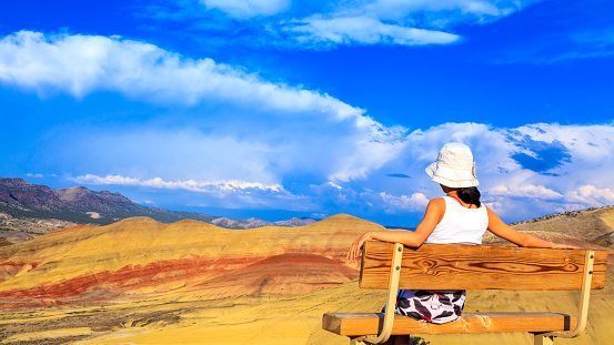 Young woman sitting on the hill-top bench relaxing while watching the colorful Painted hills at John Day Fossil Beds National Monument, Oregon