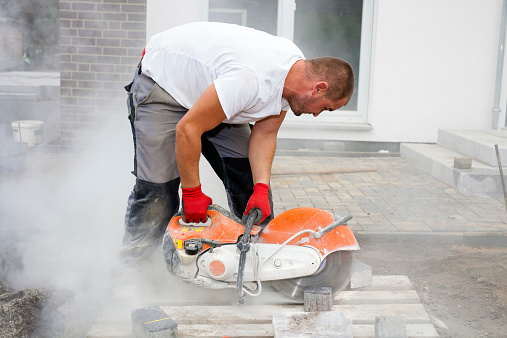 Construction worker using a concrete saw, cutting stones in a cloud of concrete dust for creating a track.