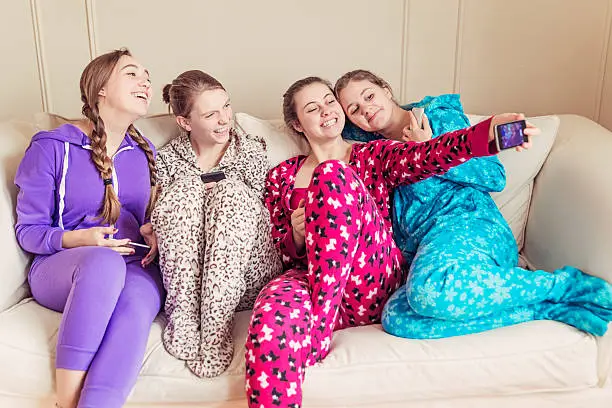 Four cute girlfriends wearing onesies chilling on a couch with mobile phones. They are laughing making selfies. Horizontal.