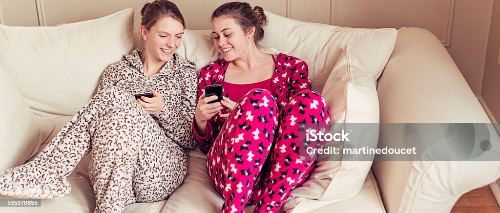 Girlfriends wearing onesies chilling on a couch with mobile phones. Two cute girlfriends wearing onesies chilling on a couch with mobile phones. Letterbox format. Infant Bodysuit Stock Photo