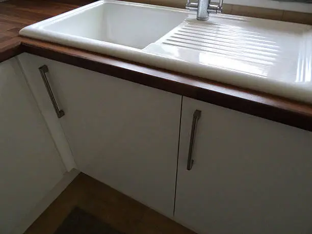 Photo showing a modern white ceramic kitchen sink and drainer / draining board with a brushed stainless-steel mixer tap.  The sink is part of a contemporary kitchen with a dark wood effect laminate worktop, and is positioned in front of a white UPVC double glazed window / windowsill, on a corner.