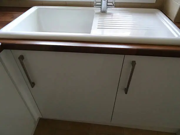 Photo showing a modern white ceramic kitchen sink and drainer / draining board with a brushed stainless-steel mixer tap.  The sink is part of a contemporary kitchen with a dark wood effect laminate worktop, and is positioned in front of a white UPVC double glazed window / windowsill.