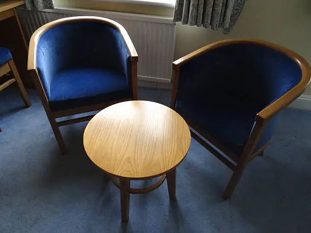 Photo showing two blue cushioned, wooden curved armchairs, pictured around a small round oak wood table, where the chairs are used as a place to sit and enjoy a cup of coffee or tea.  The chairs are often described as being semi-circular, curved, curving or banana shaped, or bucket seats, being located on one side of a bedroom.