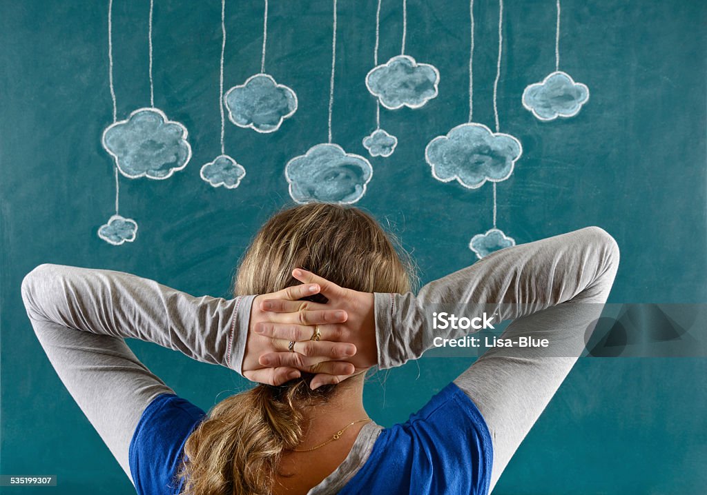 Cloud Computing Concept Pensive young woman with her face in front of a blue blackboard. She is sitting with hands behind head and looking multiple clouds. The chalkboard drawing - cloud computing concept - shows multiple clouds outlined in white and blue on a blue background. There are ten blue clouds of various sizes dangling from white strings attached to the top of the illustration. The blue background looks like a sky. 2015 Stock Photo