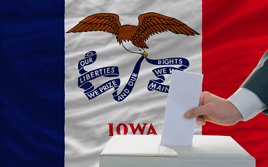 man putting ballot in a box during elections  in front of flag american state of iowa