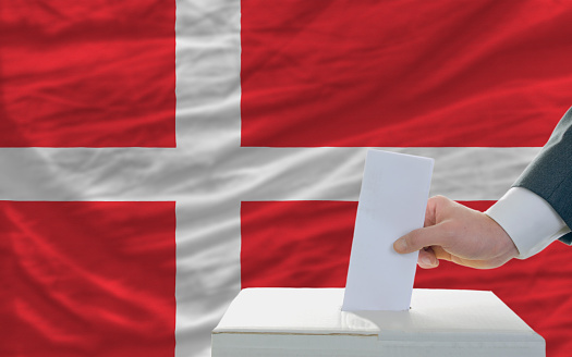 man putting ballot in a box during elections in denmark in fornt of flag