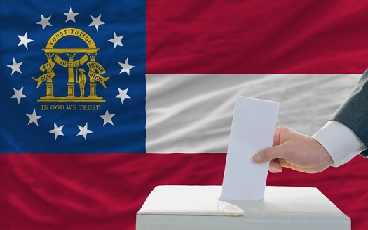 man putting ballot in a box during elections in georgia in fornt of flag