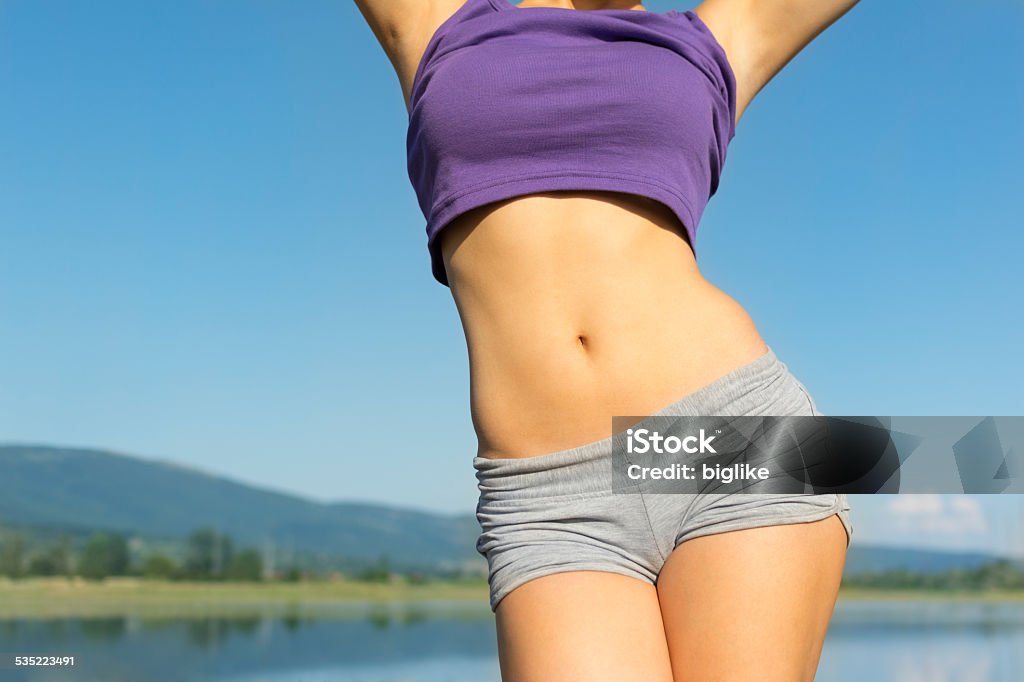 Young fit woman's belly outdoors in summer Closeup of young fit woman's belly outdoors in summer. Fitness girl in gray shorts and purple tank top outdoors at the beach on sunny summer day. No retouch, developed from RAW, vibrant colors, natural light. Women Stock Photo