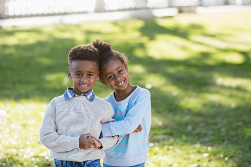 Cute little African American brother and sister standing arm in arm, smiling at the camera.  They are twins, 6 years old.  It is a bright, sunny day at the park in early spring or autum.