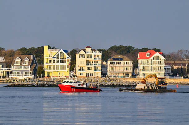 A tow boat, the American Titan out of West Ocean City, pulls a barge with an excavator and a load of cement water pipe in front of some nice waterfront properties at sunrise.