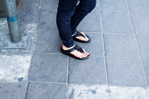 Feet of Thai woman in black sandals. Woman is wearing black jeans and is standing on sidewalk.